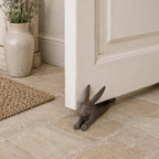 A rustic rabbit-shaped cast iron doorstop props open a white paneled door in a cozy entryway with stone flooring, a woven rug, and a decorative ceramic vase.