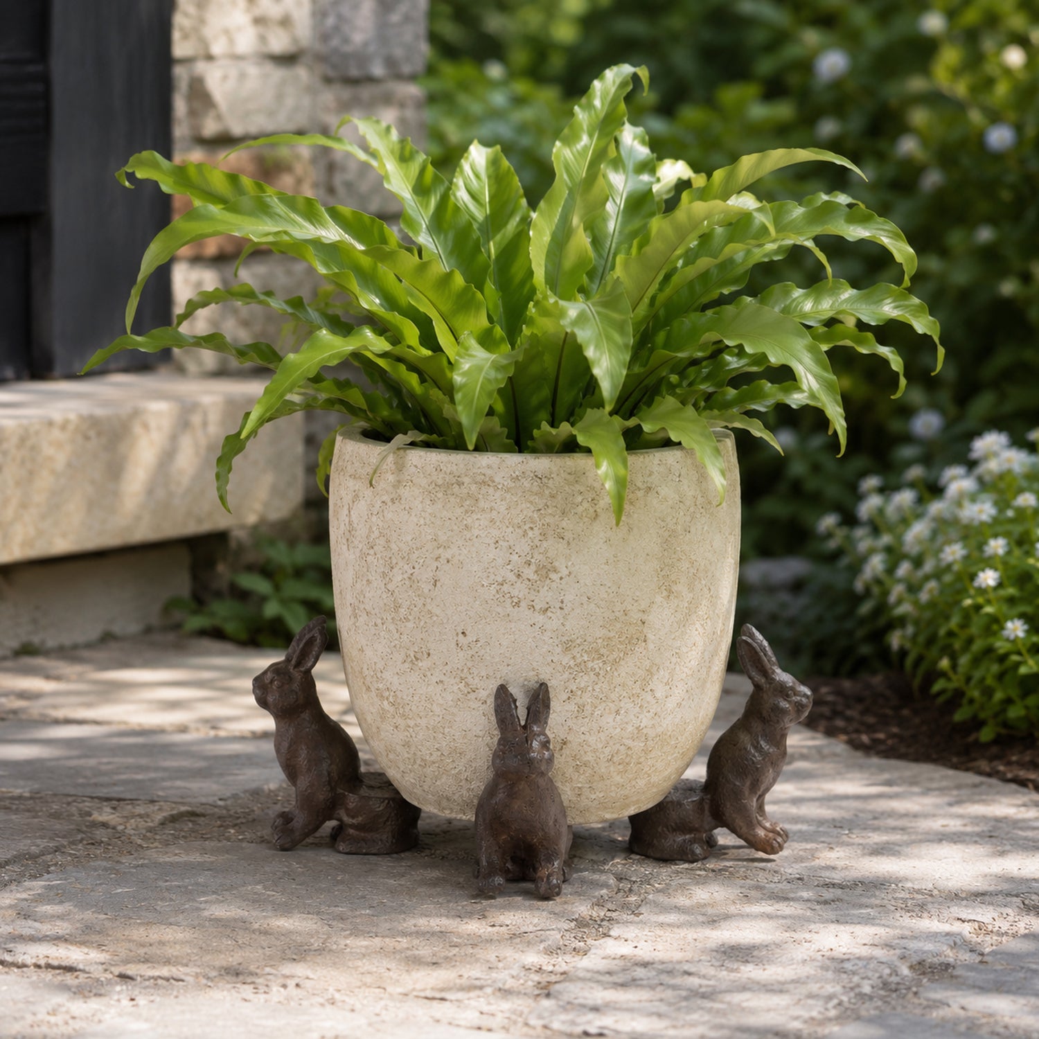 A lush green fern grows in a weathered cream-colored planter elevated by three small rabbit-shaped supports, set on a stone patio in a garden setting.