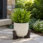 A lush green fern sits in a tall ribbed cream planter supported by three small dog-shaped feet on a stone patio beside a landscaped garden and stone house exterior.