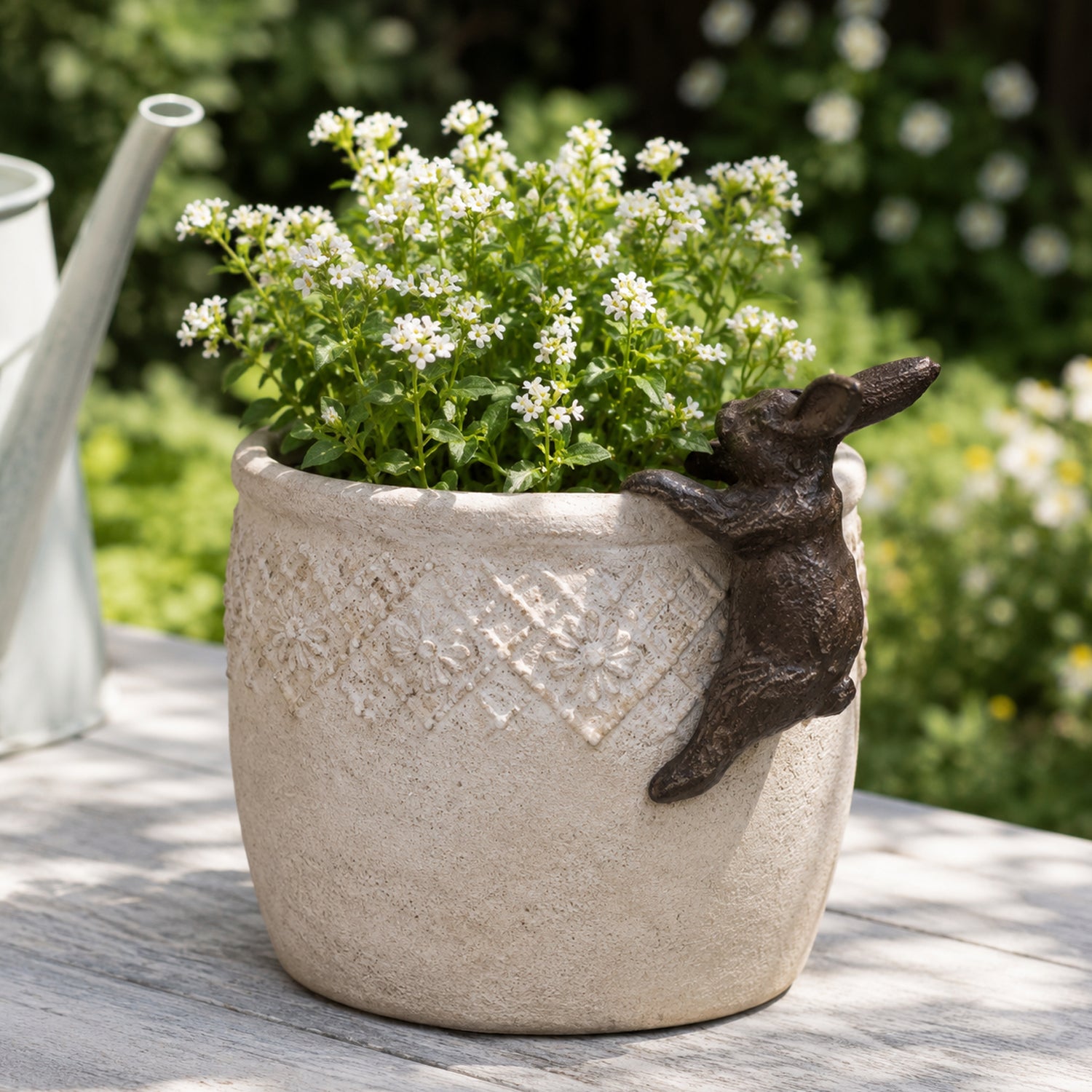 A cream-colored textured planter filled with small white flowers sits on a wooden table, decorated with a bronze-colored mouse figure climbing over its rim in a sunlit garden setting.