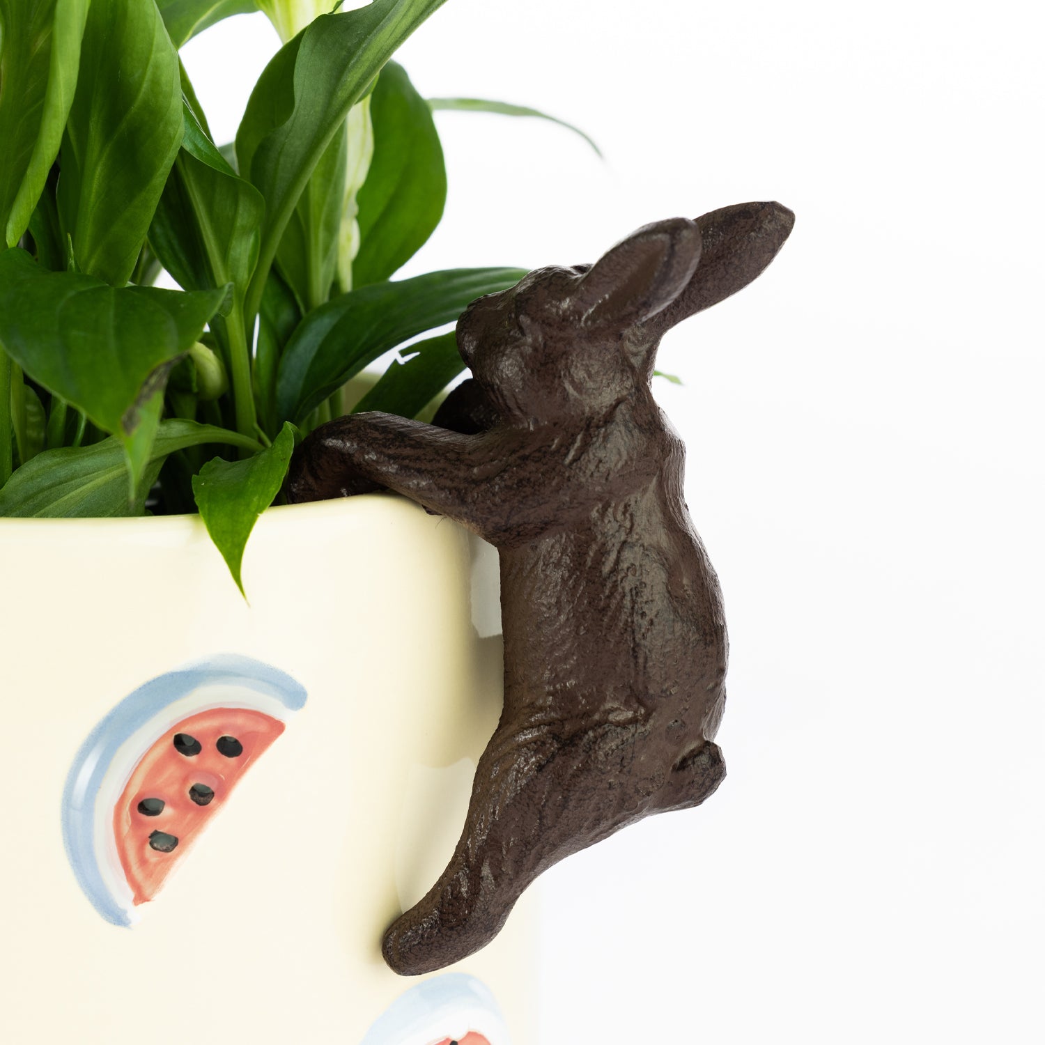 A small brown rabbit-shaped decorative accent appears to be climbing over the rim of a cream-colored planter filled with green foliage, which is decorated with a painted watermelon motif.