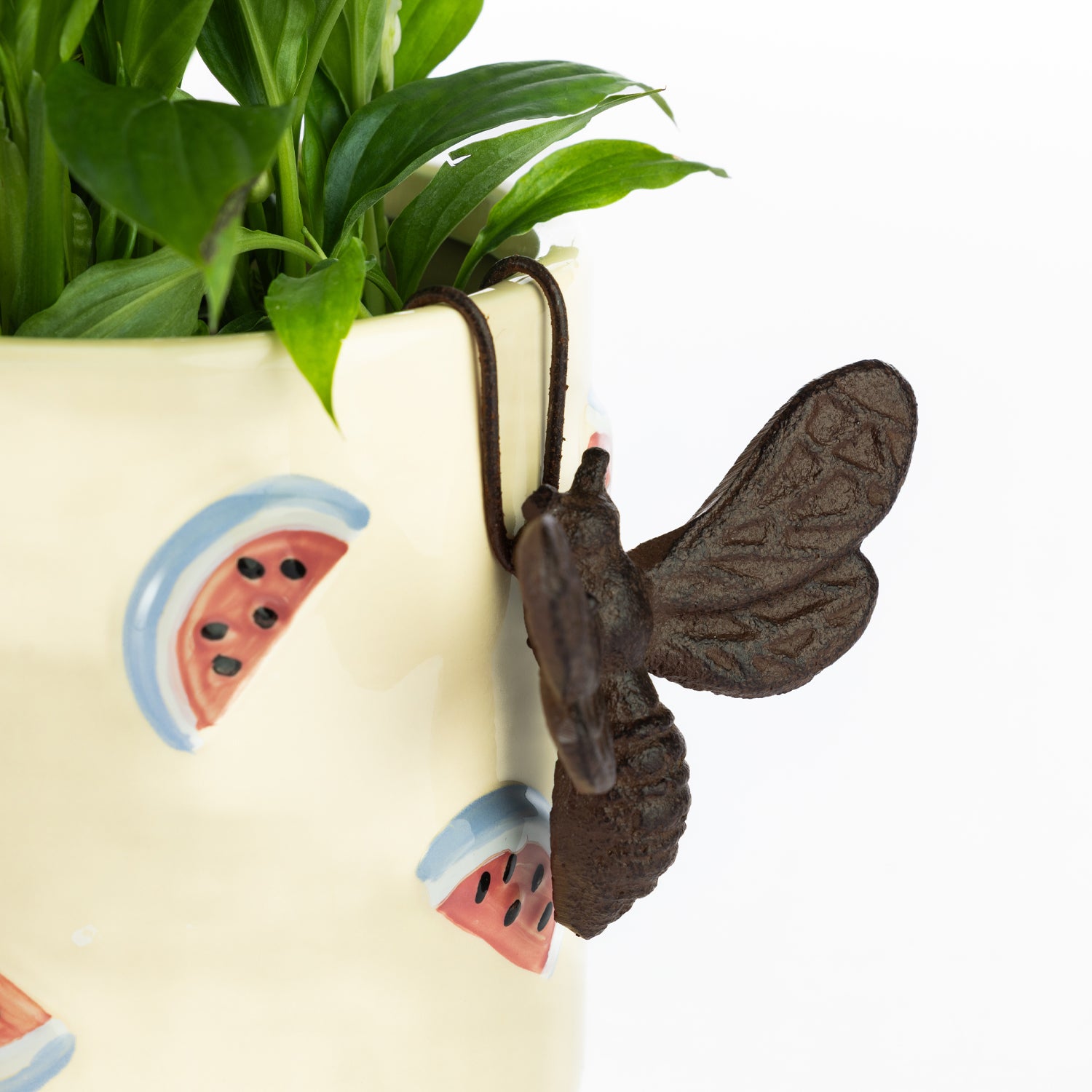 A bronze-colored bee ornament hangs from the rim of a cream-colored planter decorated with watermelon slices, beside a lush green houseplant against a plain light background.