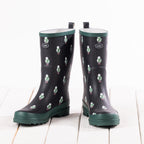 A pair of black rubber rain boots with a green and white sheep pattern, featuring dark green soles and pull tabs, displayed on a white wooden plank surface.