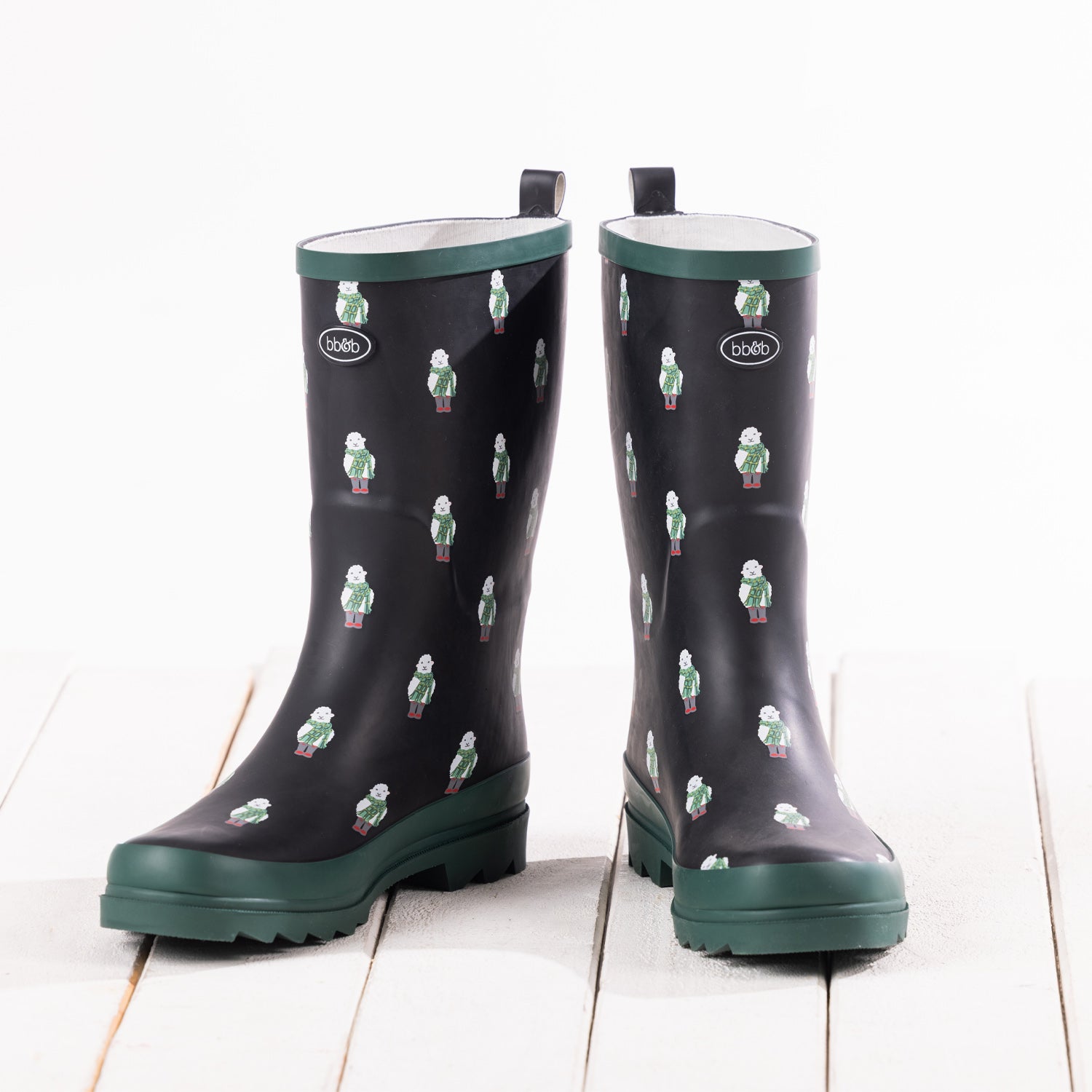 A pair of black rubber rain boots with a green and white sheep pattern, featuring dark green soles and pull tabs, displayed on a white wooden plank surface.