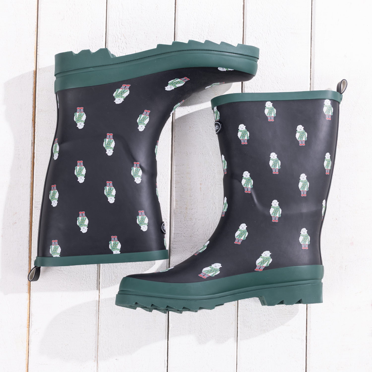 A pair of dark navy rain boots with a repeating pattern of small white and green sheep figures, featuring dark green soles and trim, displayed on a white wood plank background.