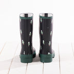 A pair of dark green rain boots with a pattern of small white and green sheep figures, shown from the back on a white wooden plank surface.