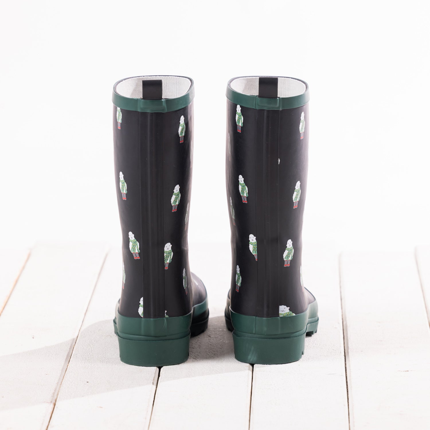 A pair of dark green rain boots with a pattern of small white and green sheep figures, shown from the back on a white wooden plank surface.