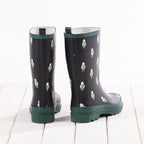 A pair of dark gray rain boots with a green and white sheep pattern, featuring solid green trim and soles, shown from the back on a white wooden plank surface.