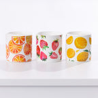 Three white ceramic candles with colorful fruit patterns, including orange slices, strawberries, and lemons, arranged on a white surface against a white background.