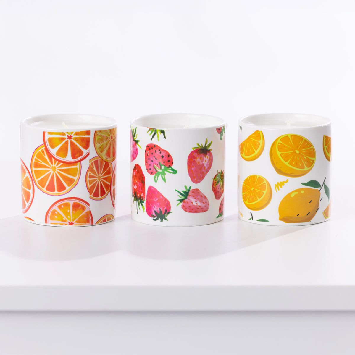 Three white ceramic candles with colorful fruit patterns, including orange slices, strawberries, and lemons, arranged on a white surface against a white background.