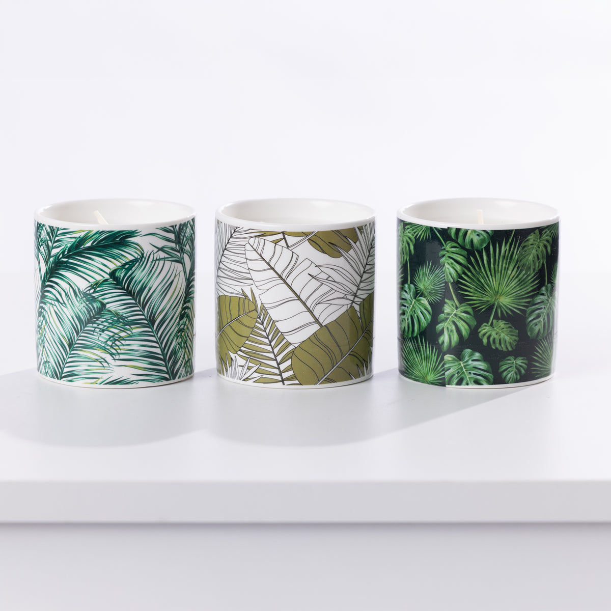 Three ceramic citronella candles with tropical leaf patterns, displayed side-by-side on a white surface against a white background. 