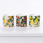 Three ceramic candles with botanical fruit and flower patterns, displayed side-by-side on a white surface against a white background.