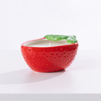 A strawberry-shaped citronella candle in a glossy red ceramic bowl with a textured seed pattern and a green leaf detail on top, set against a clean white background.