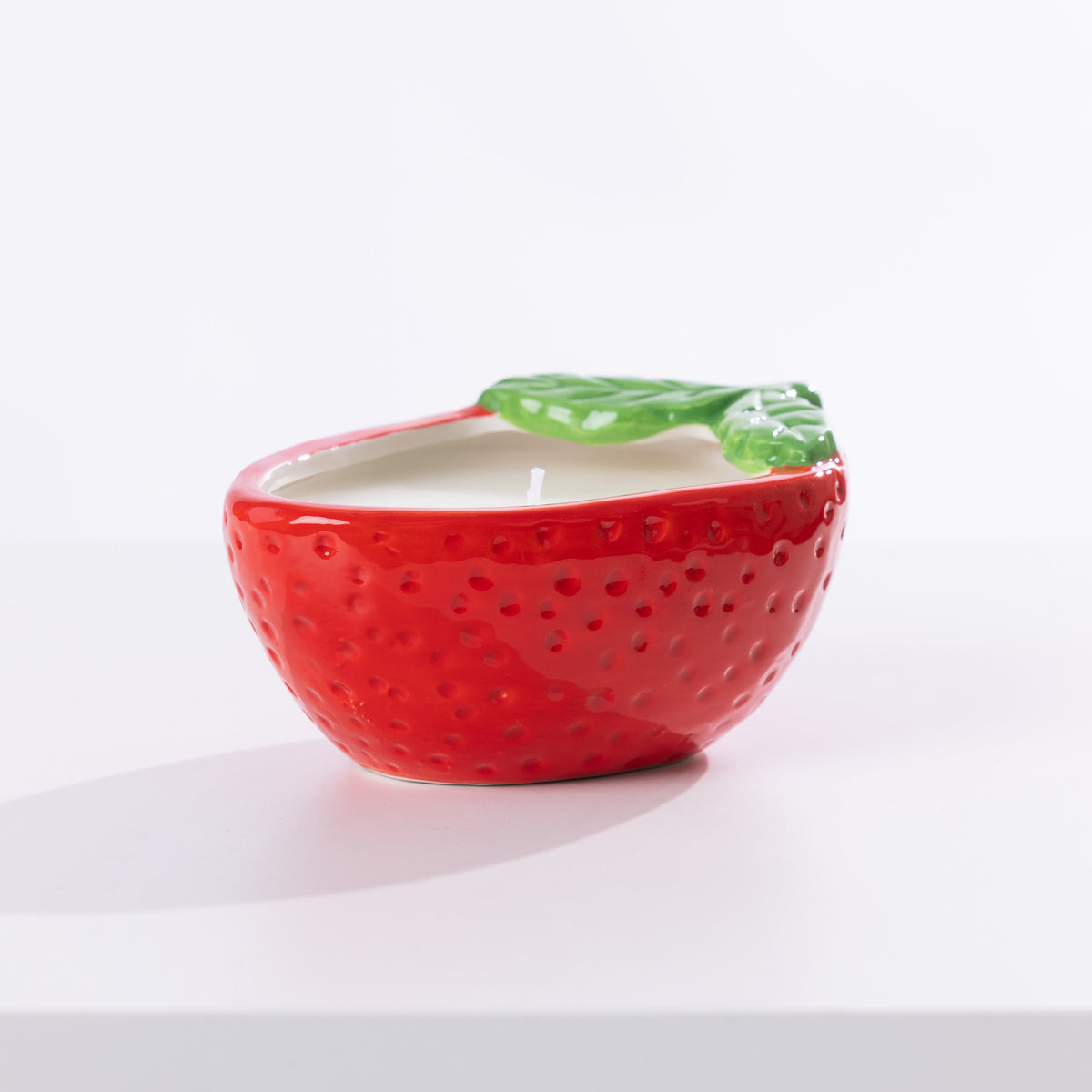 A strawberry-shaped citronella candle in a glossy red ceramic bowl with a textured seed pattern and a green leaf detail on top, set against a clean white background.