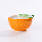An orange-shaped ceramic citronella candle with a textured orange body, a white interior, and a green leaf detail on the rim, set against a plain white background.