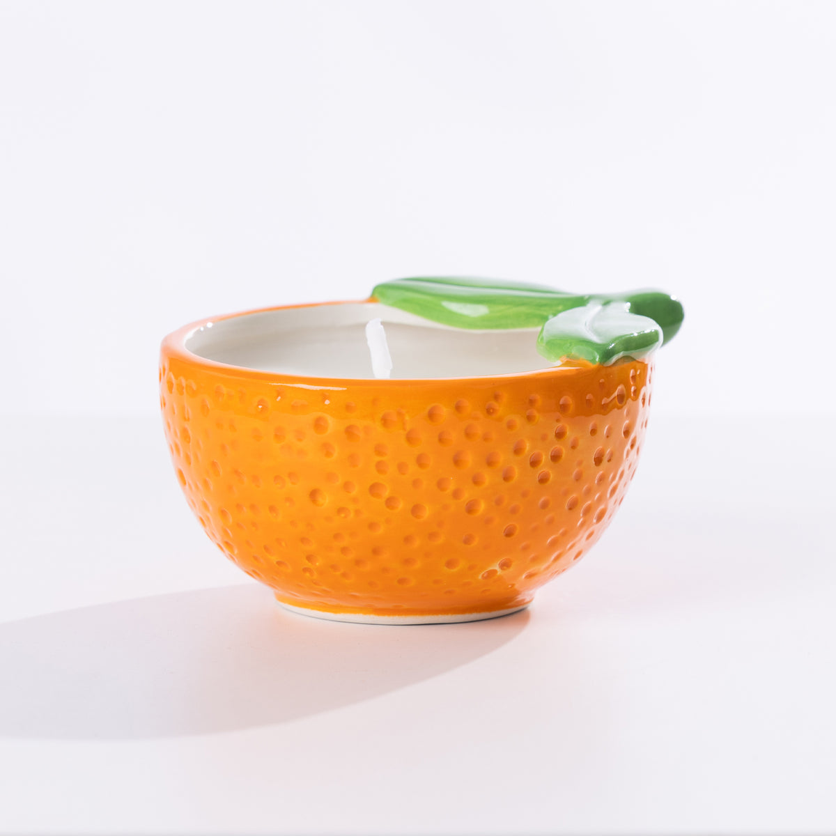 An orange-shaped ceramic citronella candle with a textured orange body, a white interior, and a green leaf detail on the rim, set against a plain white background.