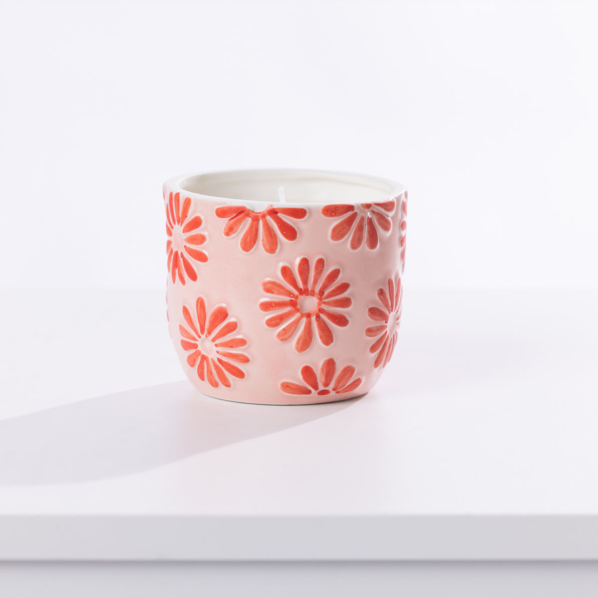 A small ceramic candle holder with a light pink body and a pattern of red daisy flowers, shown on a clean white surface with a soft shadow.