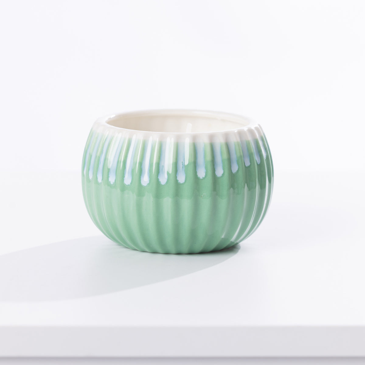 A small round ceramic candle holder with a ribbed exterior, light green glaze, and white dripping glaze accents around the rim, shown on a white surface against a white background.
