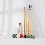 A set of croquet mallets and balls arranged on a white floor against a white wall. The set includes natural wood mallets with red, black, green, and white tips and four green, black, white, and red balls, with metal hoops.