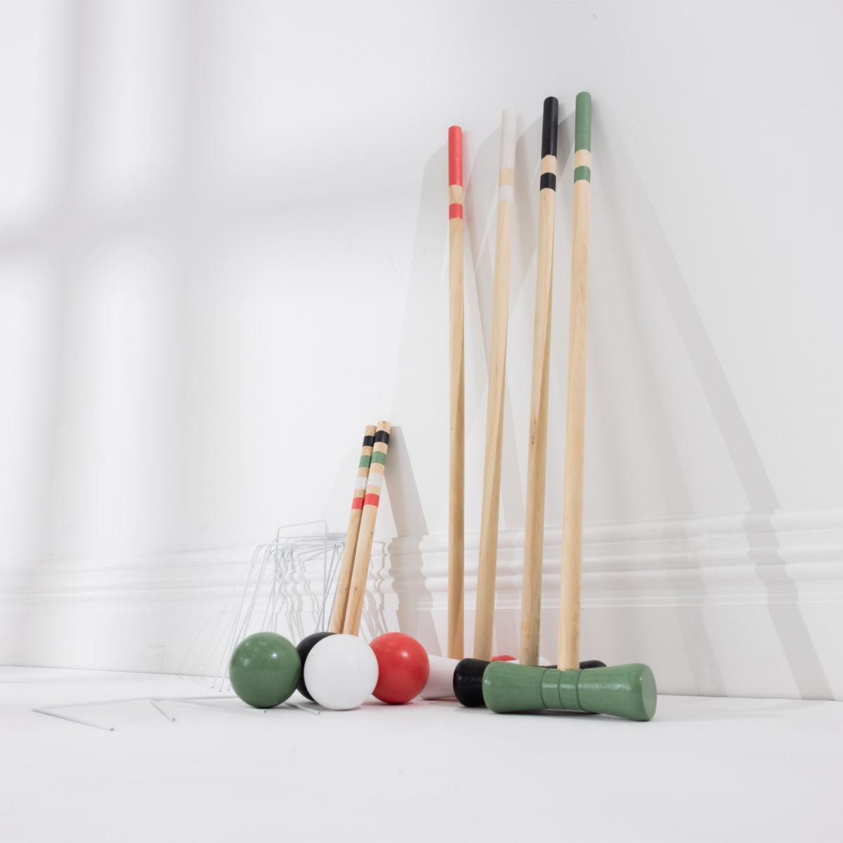 A set of croquet mallets and balls arranged on a white floor against a white wall. The set includes natural wood mallets with red, black, green, and white tips and four green, black, white, and red balls, with metal hoops.