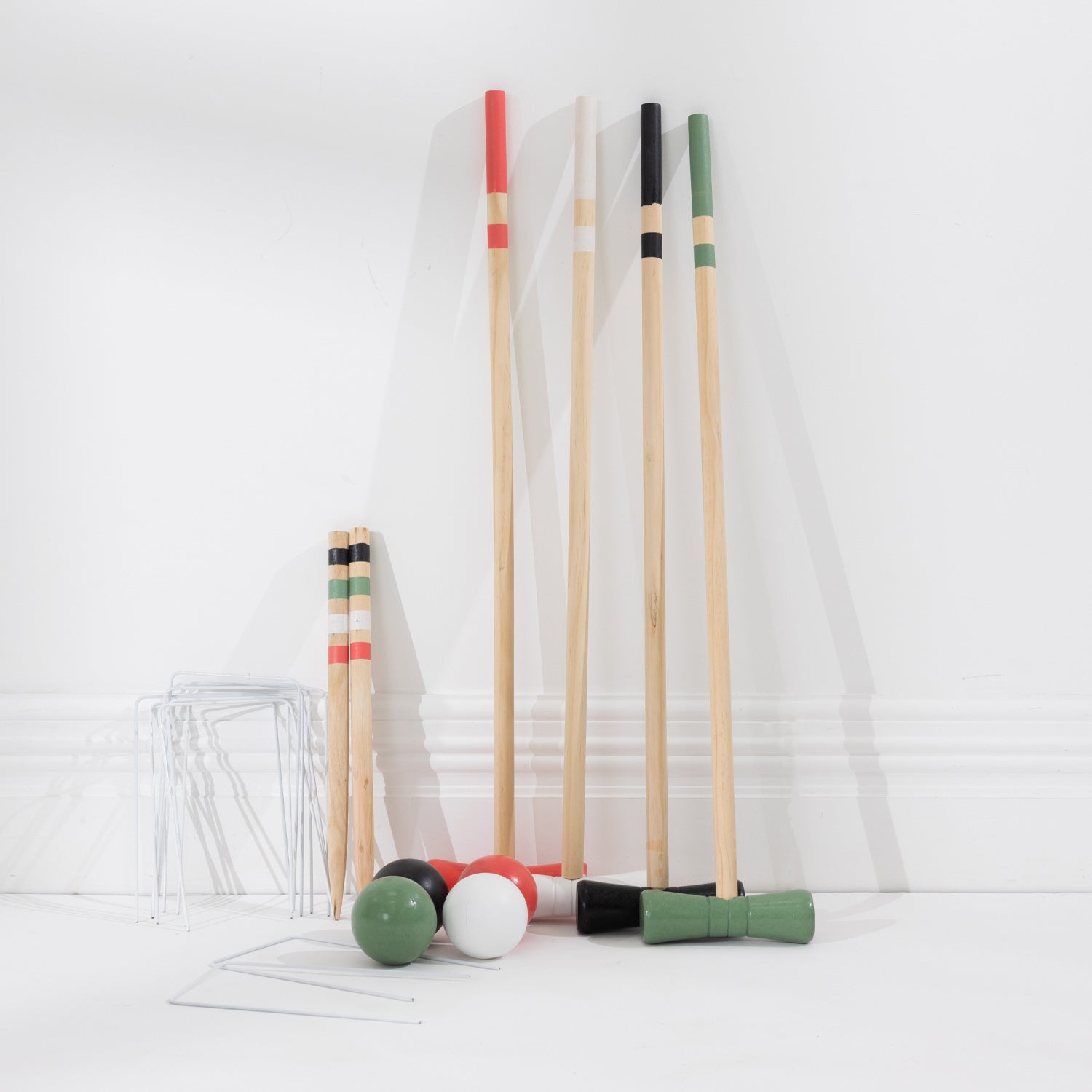 A set of croquet mallets and balls arranged on a white floor against a white wall. The set includes natural wood mallets with red, black, green, and white tips and four green, black, white, and red balls, with metal hoops.