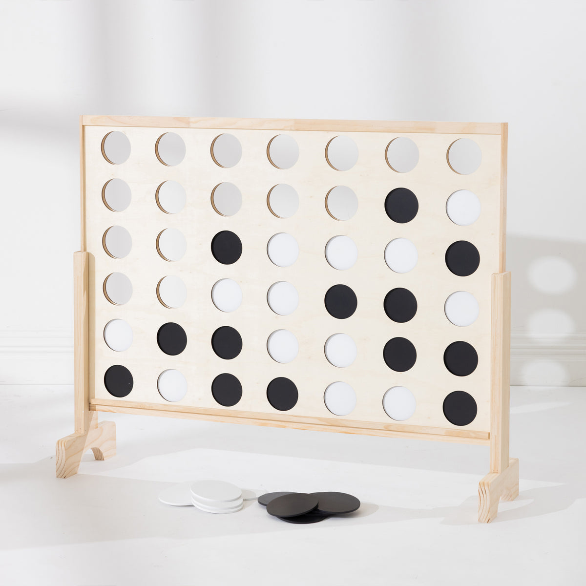 A wooden Connect-four in a row game with a light natural wood frame and black-and-white circular tokens, photographed on a clean white floor against a white wall.