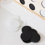 White and black round wooden discs arranged on a white surface, with a partial light wood board visible in the upper right containing circular cutouts.