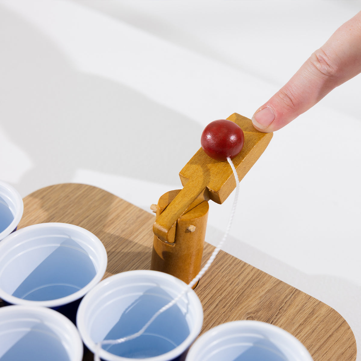 Close-up of a finger pressing the wooden launcher with a tethered red ball beside the blue cups.