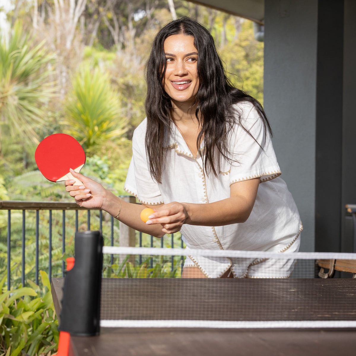 Woman holding a red table tennis paddle and orange ball while playing with the Play the Field Table Tennis Set outdoors.
