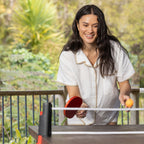 Woman playing table tennis outdoors using the Play the Field Table Tennis Set with a red paddle and orange ball.