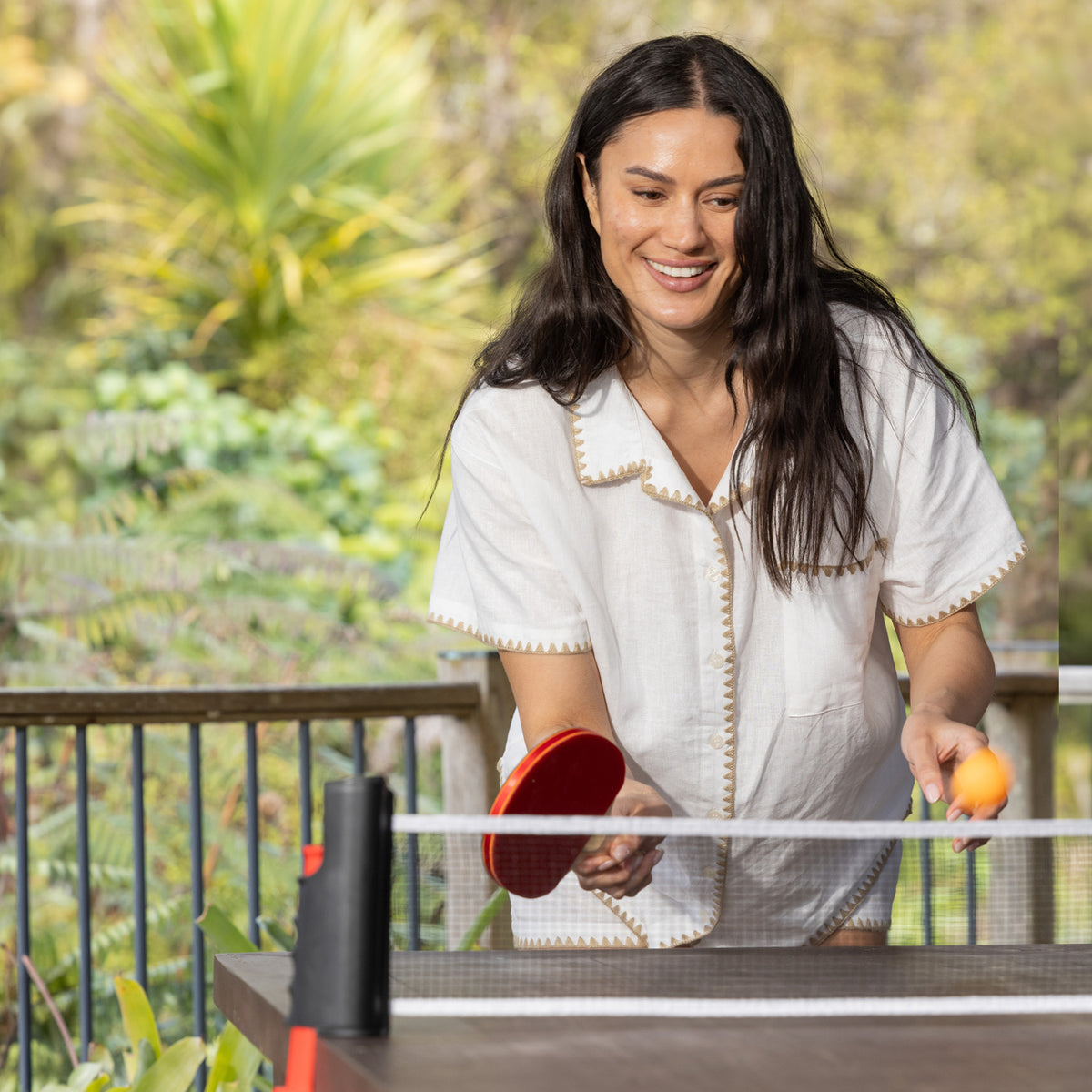 Woman playing table tennis outdoors using the Play the Field Table Tennis Set with a red paddle and orange ball.