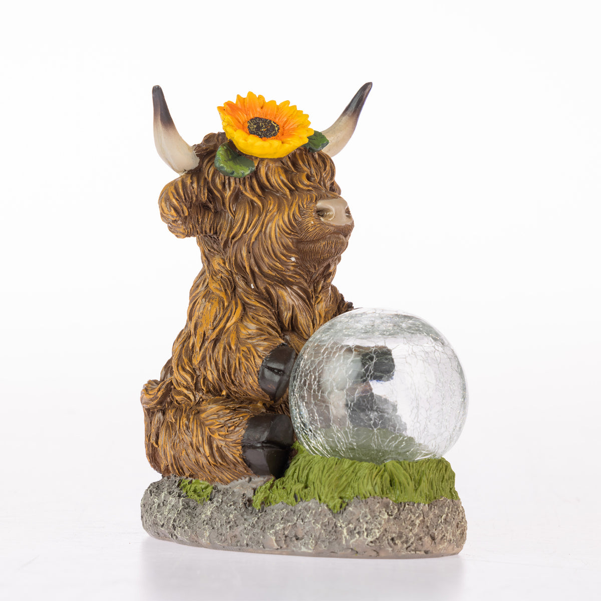 A decorative Highland cow figurine with brown shaggy fur, curved horns, a yellow sunflower on its head, and a clear cracked-glass orb resting on green grass atop a stone base.