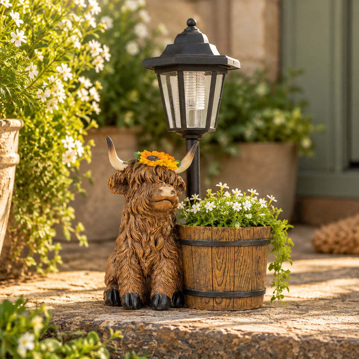 A decorative outdoor scene featuring a brown highland cow figurine with a yellow sunflower crown beside a wooden planter and a black lantern on a stone path.