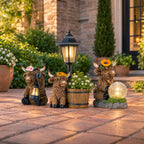 Three decorative Highland cow garden statues with sunflower crowns and lanterns are arranged on a stone patio in front of a house, alongside a wooden planter and a glowing solar globe light.