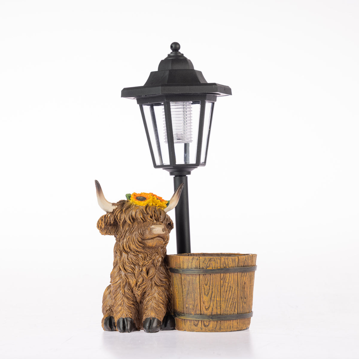 A decorative garden scene featuring a brown Highland cow figurine with a yellow flower crown, next to a black street lamp and a small wooden barrel planter, set against a plain white background.