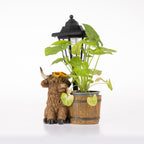 A decorative garden planter featuring a brown highland cow figurine with a yellow flower on its forehead, next to a small wooden barrel planter holding a green leafy plant, with a black lantern behind it, all on a white background.