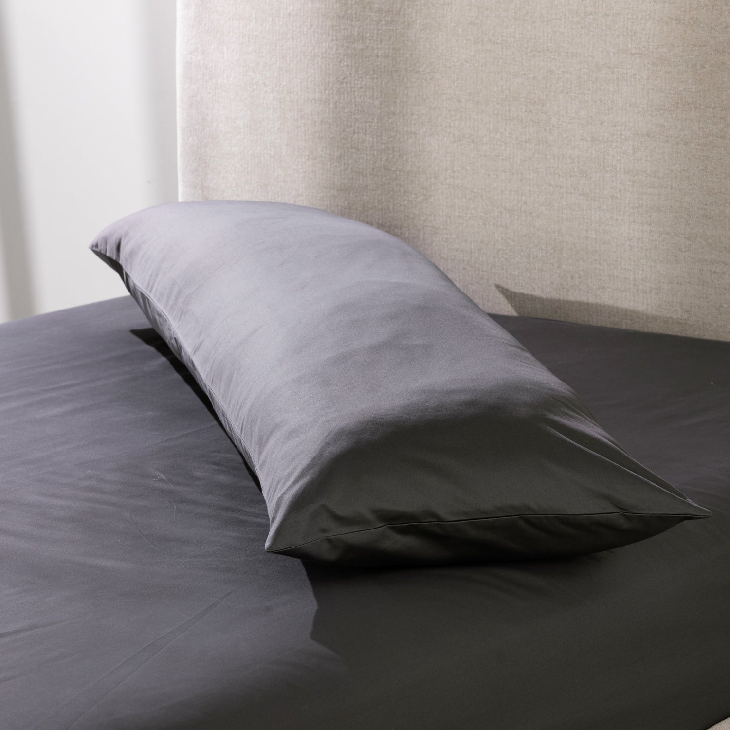 A charcoal body pillow is dressed in a smooth cotton pillowcase and styled on a neatly made bed, showcasing its clean, luxurious finish and full-length comfort.