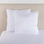 White satin Euro pillowcase pair styled on a bed against a neutral upholstered headboard.