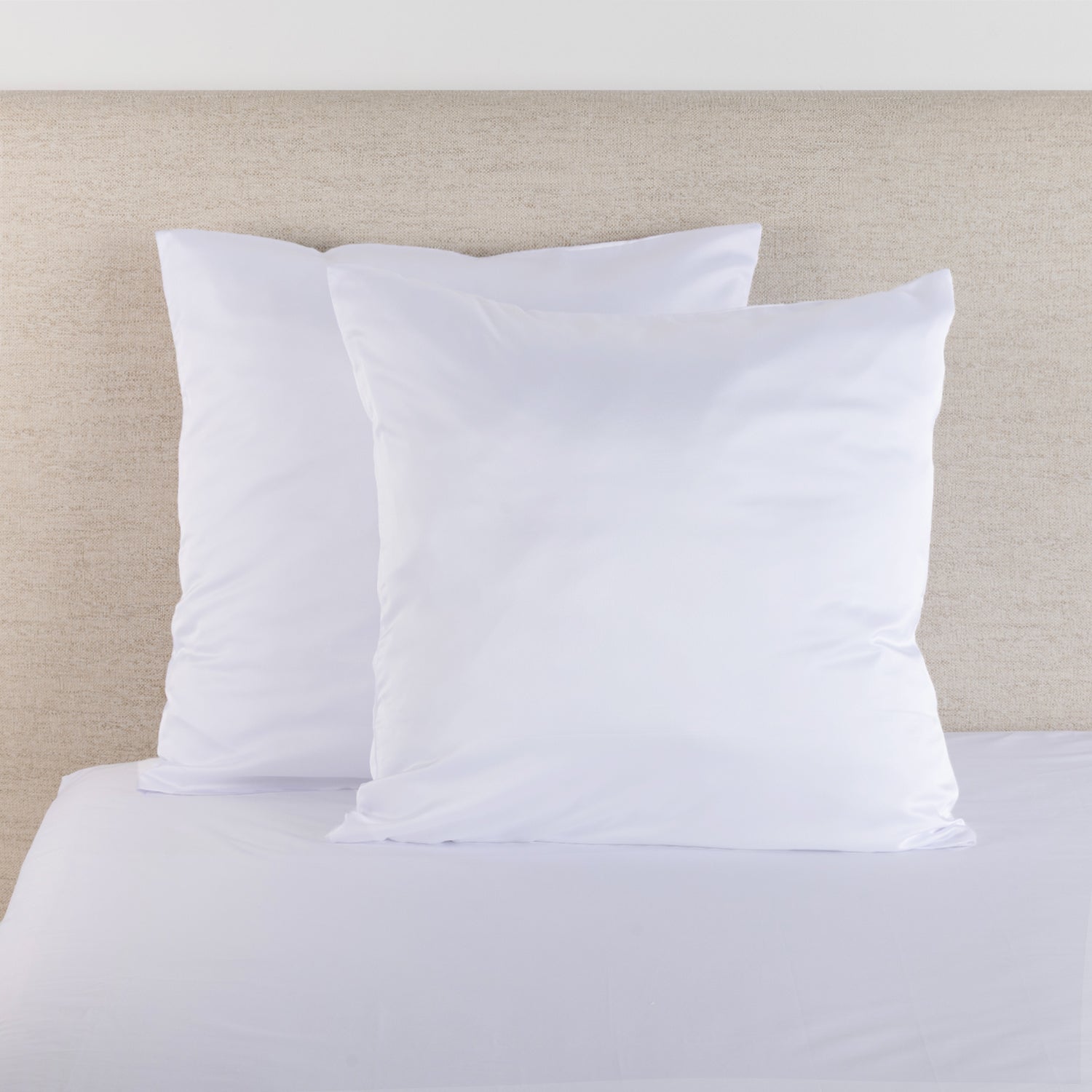 White satin Euro pillowcase pair styled on a bed against a neutral upholstered headboard.