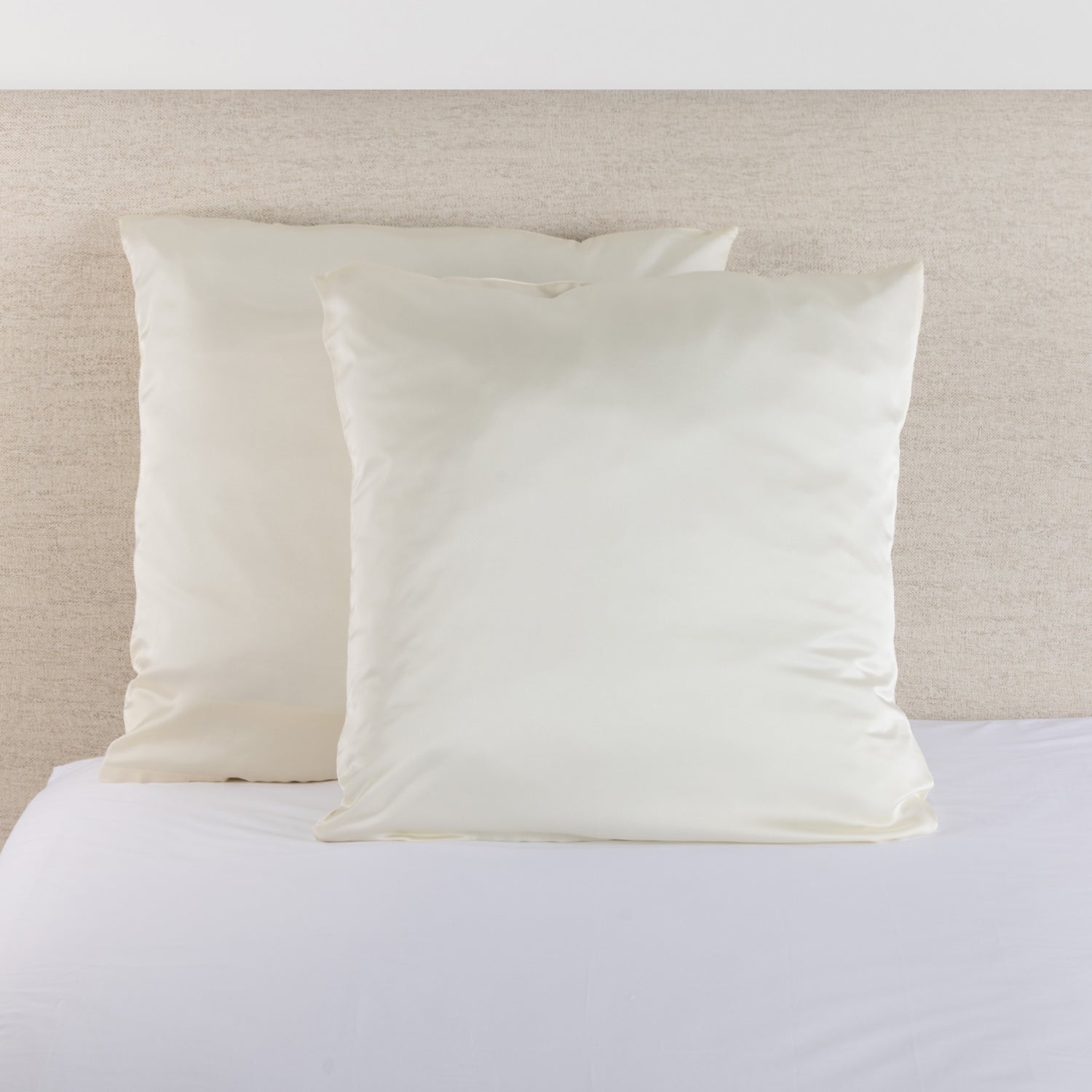 Cream satin Euro pillowcase pair styled on a bed against a neutral upholstered headboard.