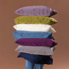 A tall stack of colorful satin pillows in lavender, olive green, ivory, blue, purple, gray, and navy is balanced in front of a person against a warm brown studio backdrop.