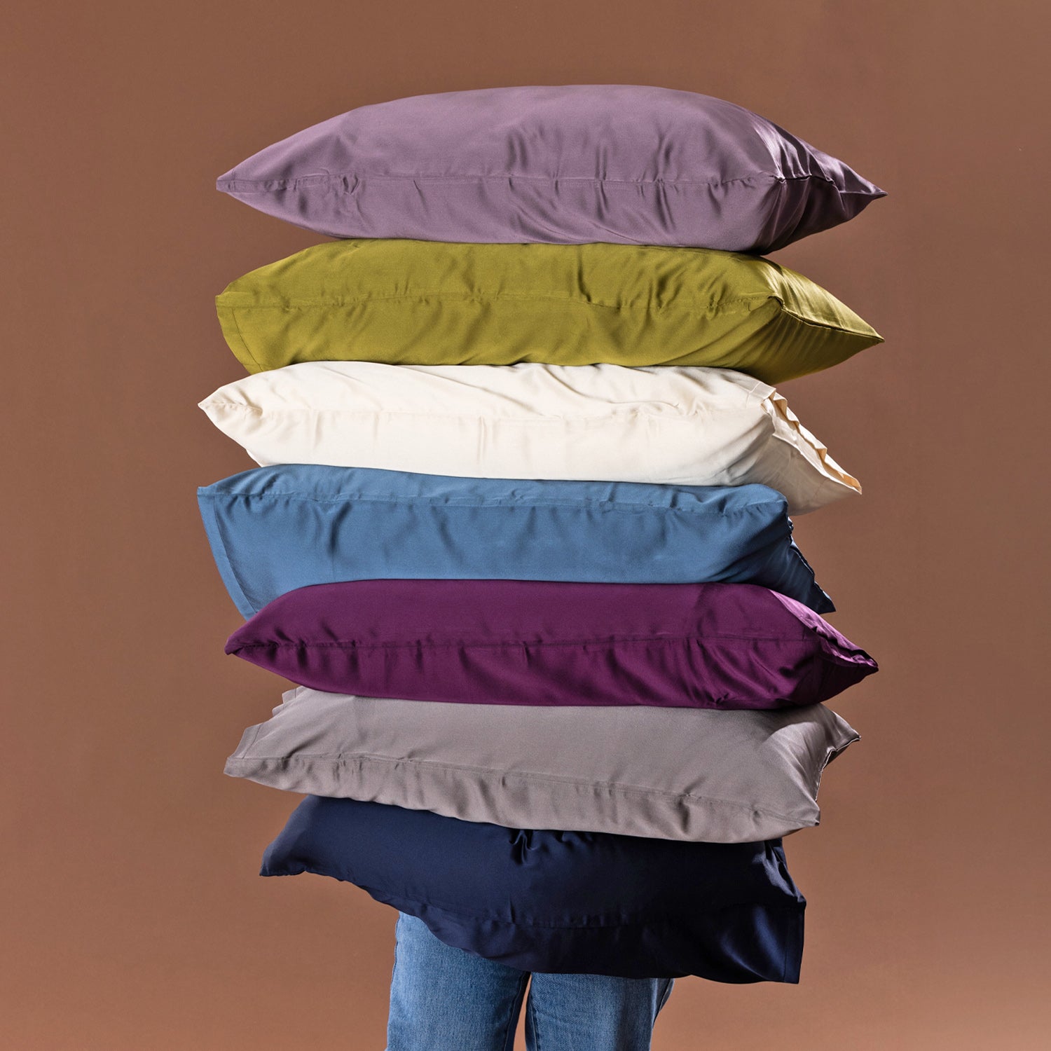 A tall stack of colorful satin pillows in lavender, olive green, ivory, blue, purple, gray, and navy is balanced in front of a person against a warm brown studio backdrop.