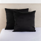 Black satin Euro pillowcase pair styled on a bed against a neutral upholstered headboard.