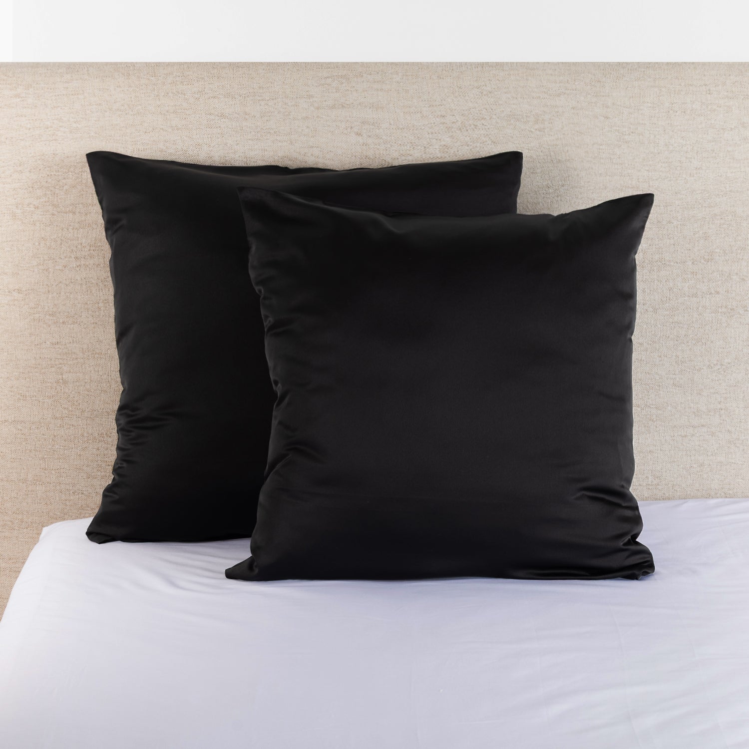 Black satin Euro pillowcase pair styled on a bed against a neutral upholstered headboard.