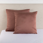 Dusky pink satin Euro pillowcase pair styled on a bed against a neutral upholstered headboard.