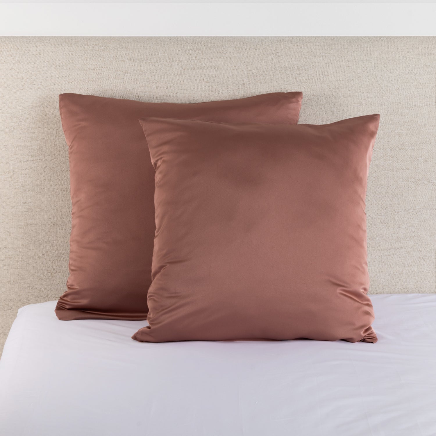 Dusky pink satin Euro pillowcase pair styled on a bed against a neutral upholstered headboard.