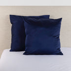 Midnight navy satin Euro pillowcase pair styled on a bed against a neutral upholstered headboard.