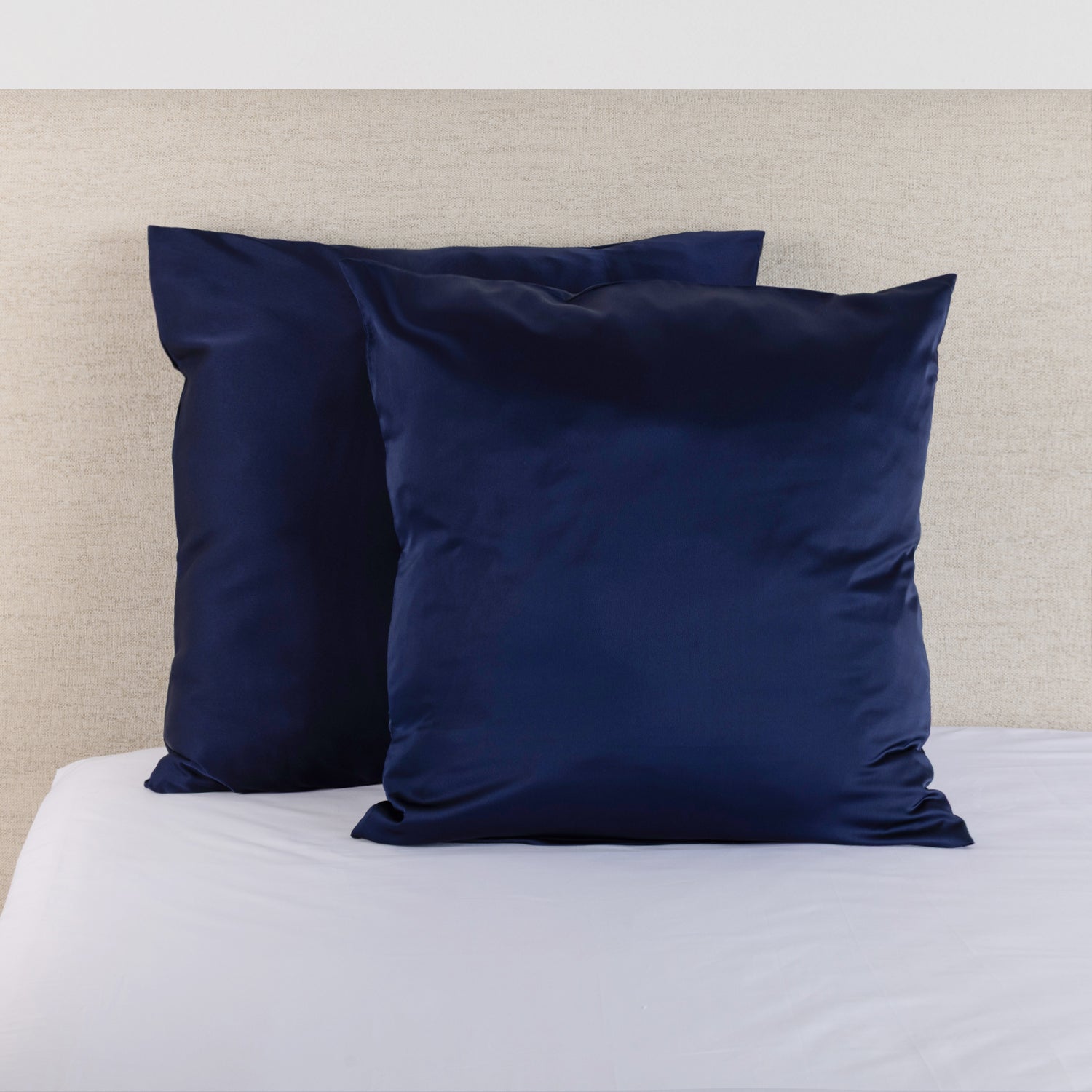 Midnight navy satin Euro pillowcase pair styled on a bed against a neutral upholstered headboard.