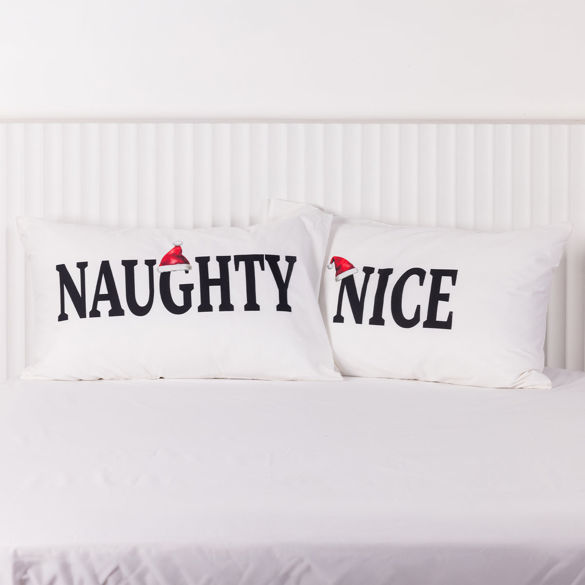 Christmas Wishes Naughty and Nice Pillowcase Pair displayed side by side on a white bed, featuring black lettering and red Santa hats.