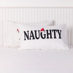 White Christmas Wishes Naughty and Nice pillowcases featuring bold black lettering and festive Santa hats, displayed overlapping on a white bed.